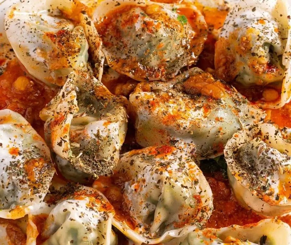 Mantu Teigtaschen mit Fleischfüllung in Tomatensauce, serviert mit Joghurt und getrockneten Kräutern - traditionelles zentralasiatisches Gericht